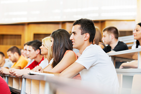 Large group of college students sitting in the university amphitheatre. The focus is on the male student in white t-shirt.
